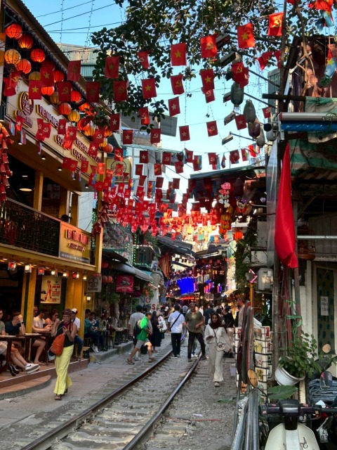 ハノイ旧市街のトレインストリートで、線路の上を歩く人々と沿道のカフェ、頭上に飾られた赤い旗とランタン。