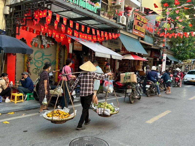 ハノイ旧市街の通りで、天秤棒を担いで果物を売り歩く行商の男性と、旗飾りが並ぶ街並み